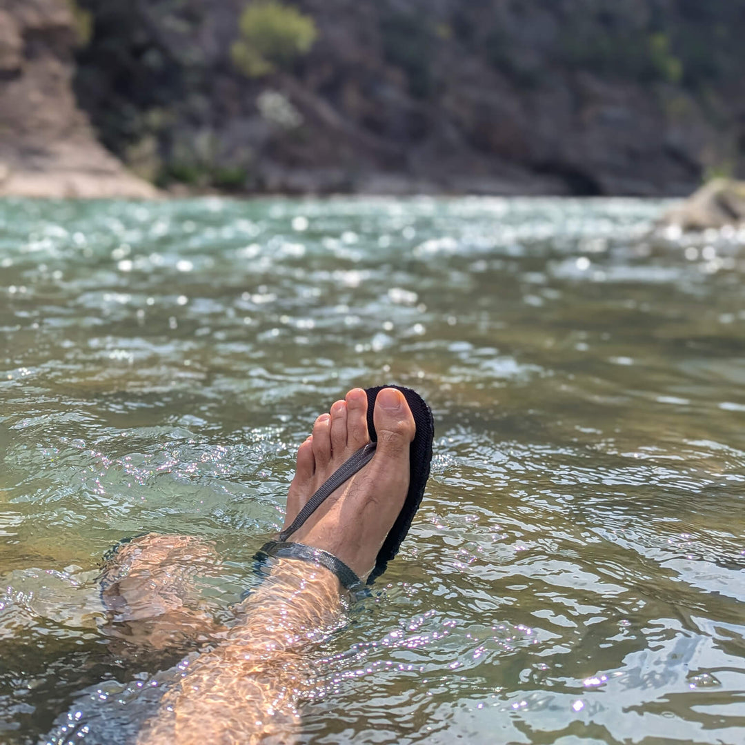 Feet in the river wearing ZuZu Flex barefoot water performance sandals with a breathable canvas footbed and replaceable recycled straps, designed for natural movement and all-day comfort.