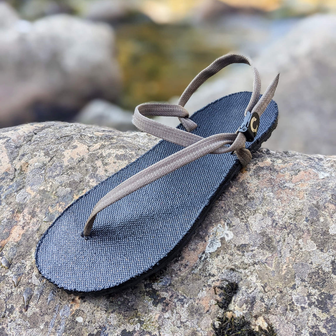 ZuZu Flex barefoot water performance sandal with a breathable canvas footbed and replaceable recycled straps, designed for natural movement and all-day comfort. On a rock in the river