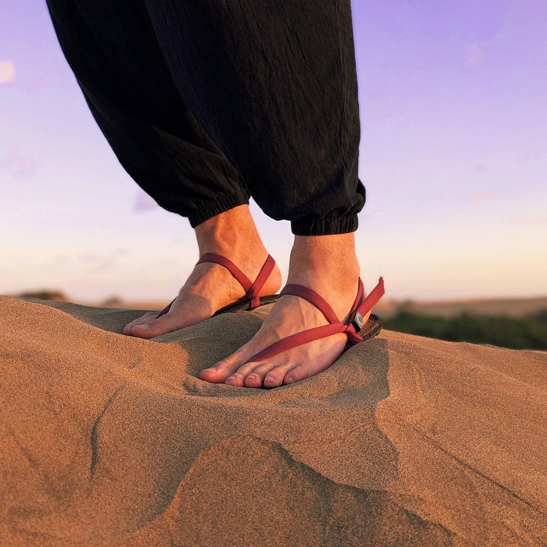 A man in black standing on a dune. Wearing ZuZu Flex barefoot sandals with a urable canvas footbed and burgundy recycled straps. Eco-friendly barefoot adventure sandals