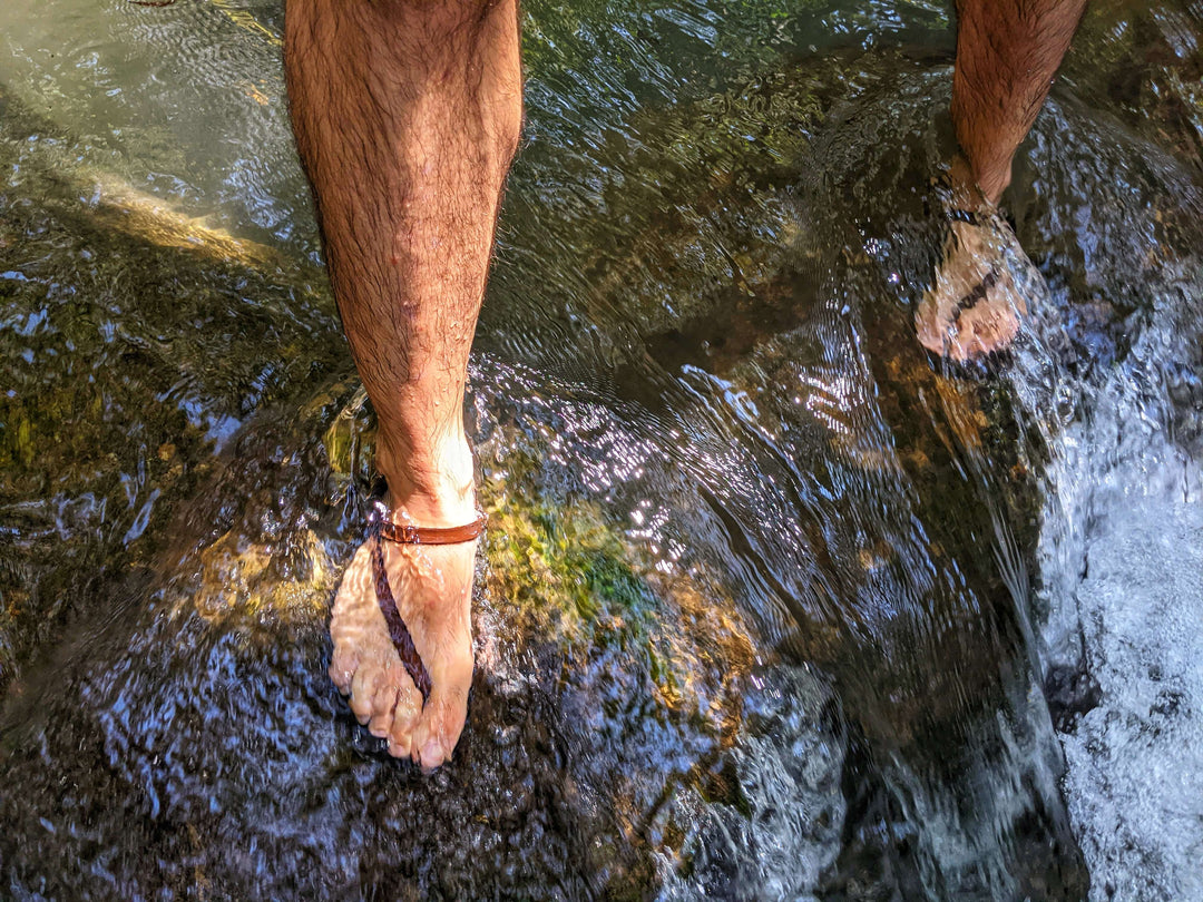 A man wearing ZuZu Flex barefoot sandals featuring a canvas footbed and replaceable leather and black color recycled strap. Standing in a river